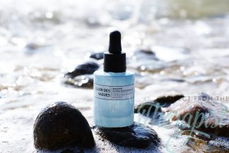 A blue dropper bottle of Des Vagues Replenishing Booster rests on wet rocks by the sea, with gentle waves and sunlight in the background.