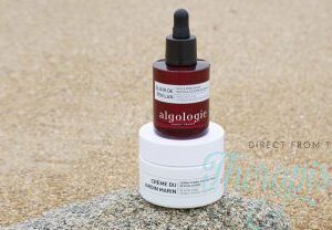 A red dropper bottle labeled Marine Gardens Precious Oil rests atop a white jar on sandy ground, both displaying minimalist white labels with text.