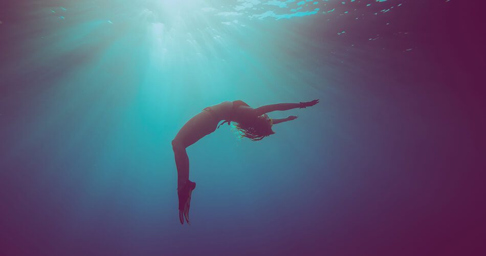 A person is gracefully arching their back underwater, arms and legs extended, with sunlight streaming down from the surface, creating a serene and ethereal scene—like a moment of tranquillity after using Algologie Skin, Home Direct From The Therapist.