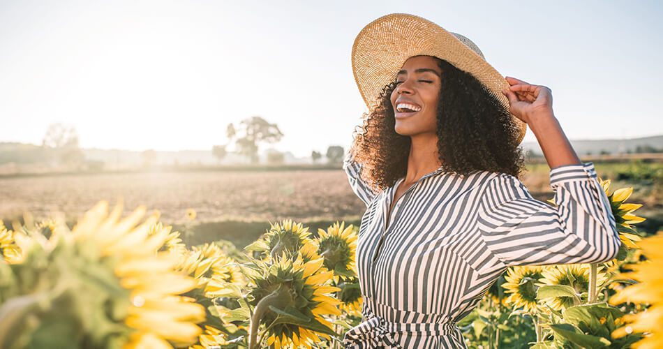 A woman wearing a striped dress and a wide-brimmed hat smiles joyfully while standing in a sunflower field on a sunny day, radiating the fresh glow of Algologie Skin care you’d expect Home Direct From The Therapist.