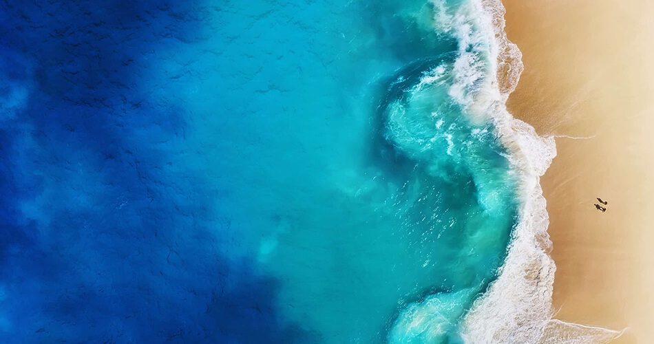 Aerial view of a sandy beach with turquoise waves crashing onto the shore. Two small figures walk near the water’s edge, where the deep blue ocean transitions to lighter blue—nature’s own Algologie Skin glow, Home Direct From The Therapist.
