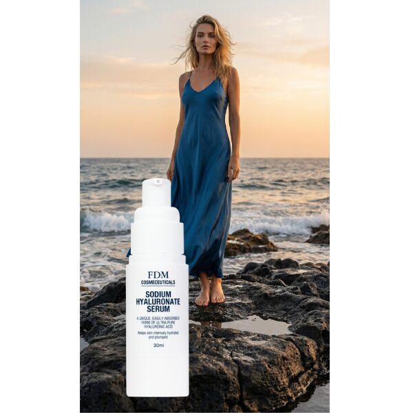 A woman in a flowing blue dress stands barefoot on rocks by the sea at sunset. In the foreground, a large bottle of FDM Cosmeceuticals Sodium Hyaluronate Serum is prominently displayed.