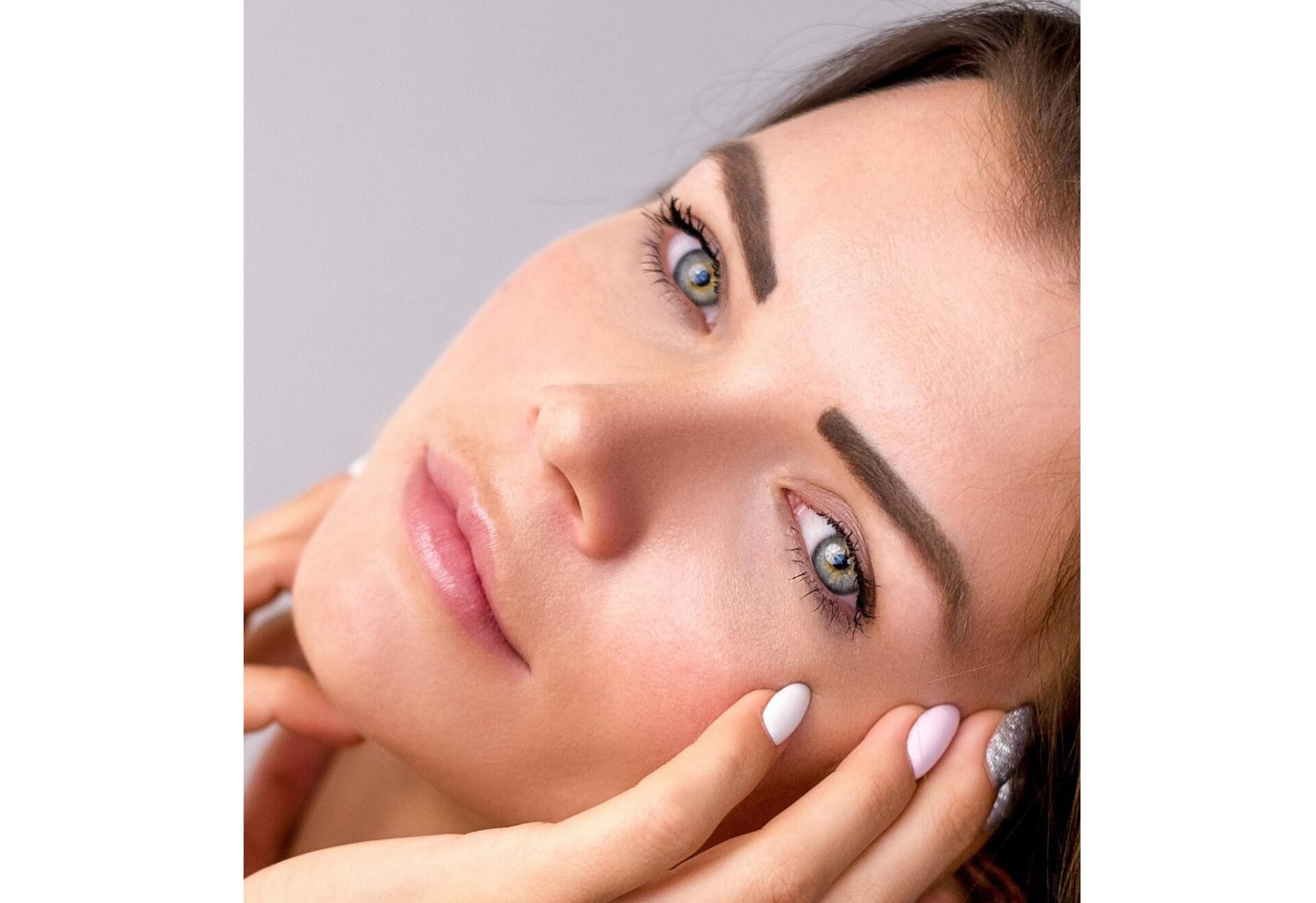 A close-up of a woman with light eyes, groomed eyebrows, and clear skin gently touching her face with manicured hands. She has a calm expression and is looking slightly upward.