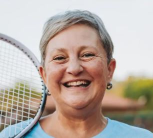 Smiling older woman with short gray hair holds a tennis racket over her shoulder outdoors, wearing a light blue shirt. The background is blurred with hints of trees and buildings.